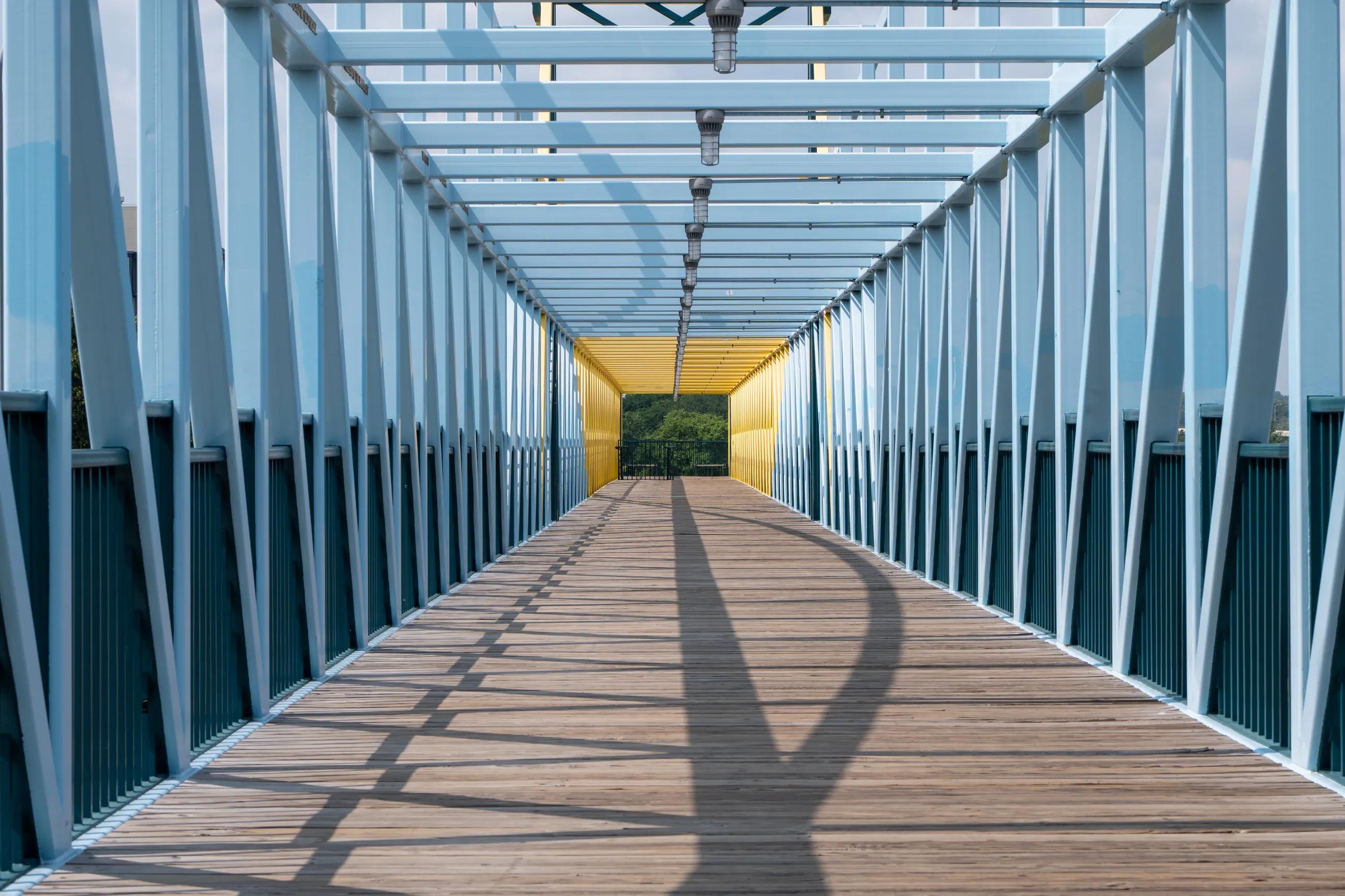Modern architectural footbridge with blue and yellow elements leading to green trees on a sunny day